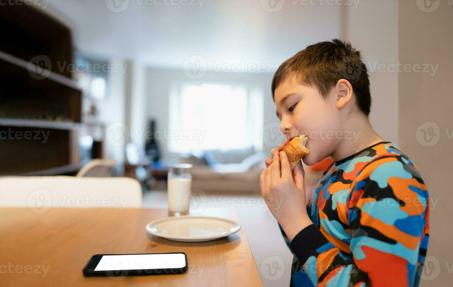 Healthy Kid having breakfast, Happy Child Boy using mobile phone watching, reading or playing games a while eating food, School Kid croissant and milk before go to school in the morning photo