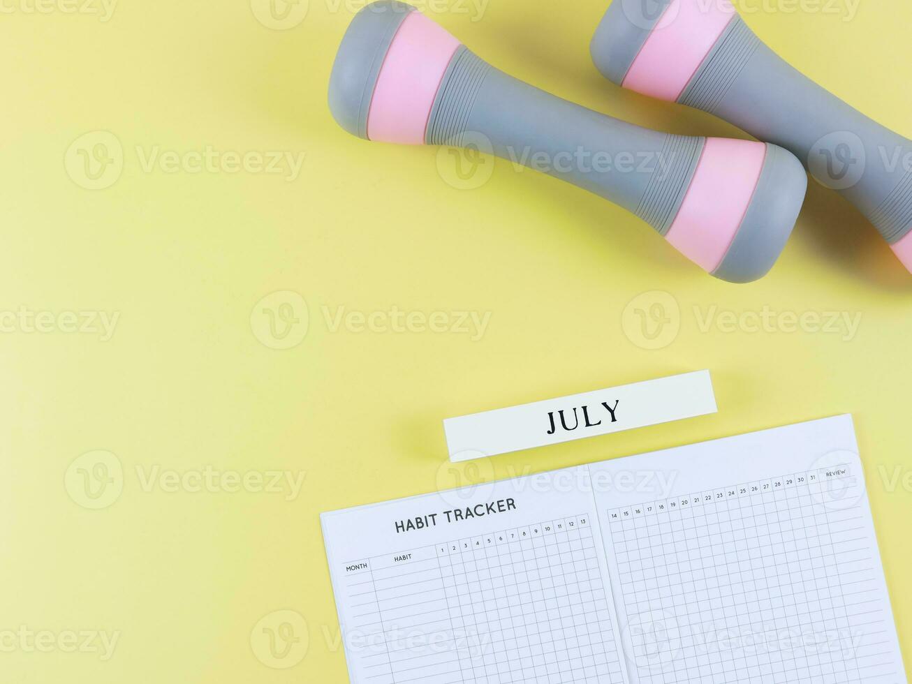 flat lay of habit tracker book, wooden calendar July, gray pink dumbbells on yellow  background with copy space. Self development and active lifestyle concept. photo