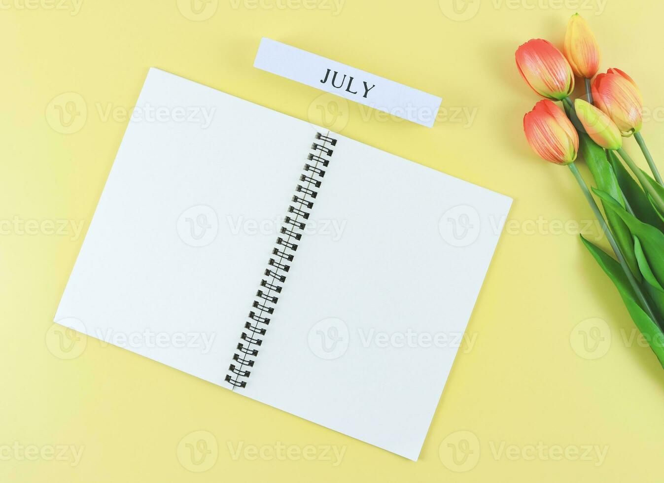 flat lay of opened blank pages notebook or diary, wooden calendar July with tulips bouquet on yellow background  with copy space. photo