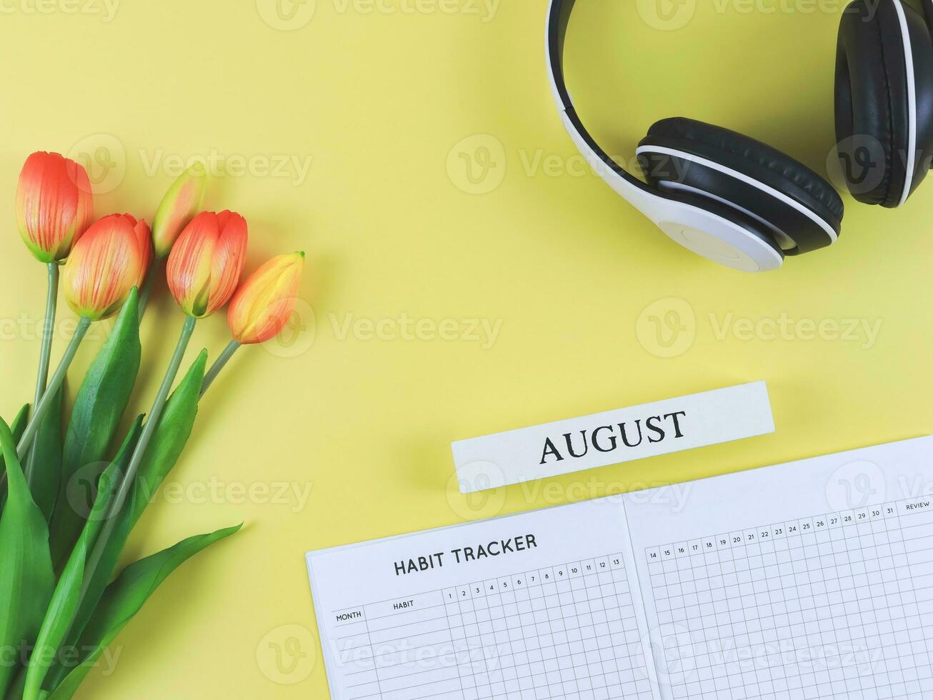flat lay of habit tracker book with wooden calendar August,  headphones and tulips  on yellow background. photo