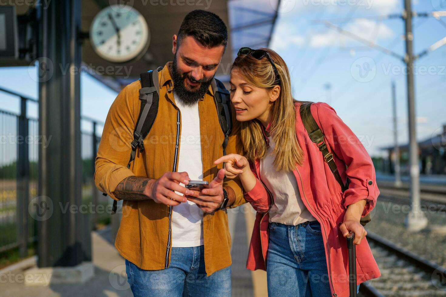 Happy couple is standing at railway station and waiting for arrival of their train. They are looking at map on smartphone. photo