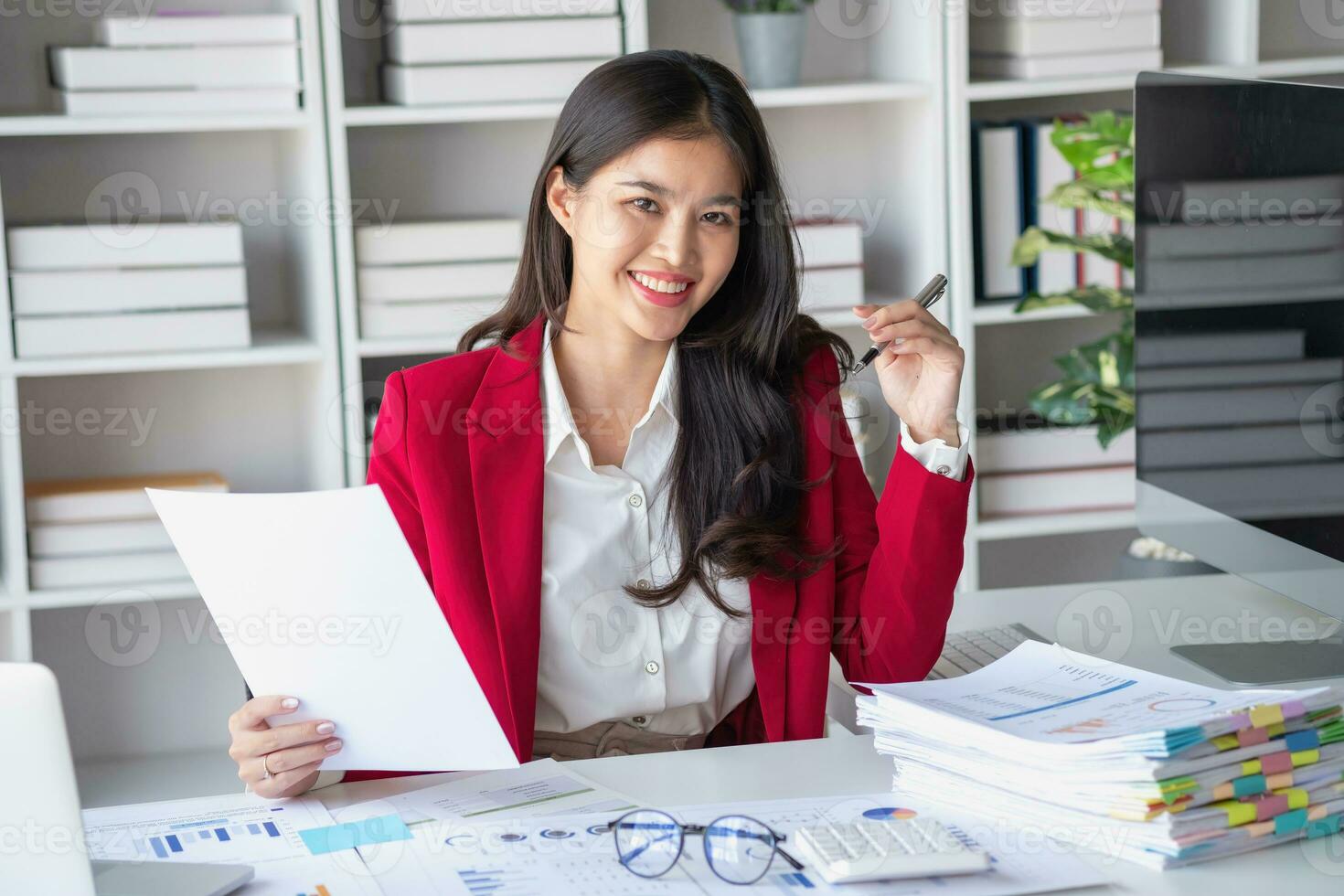Asian businesswoman in red suit calculating company sales with calculator, laptop, and tablet on table Interior of businesswoman office at the modern workstation. photo
