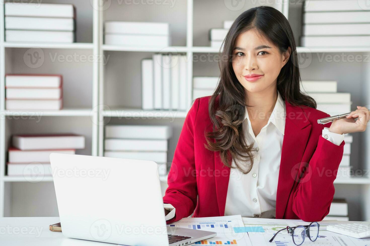 Asian businesswoman in red suit calculating company sales with calculator, laptop, and tablet on table Interior of businesswoman office at the modern workstation. photo