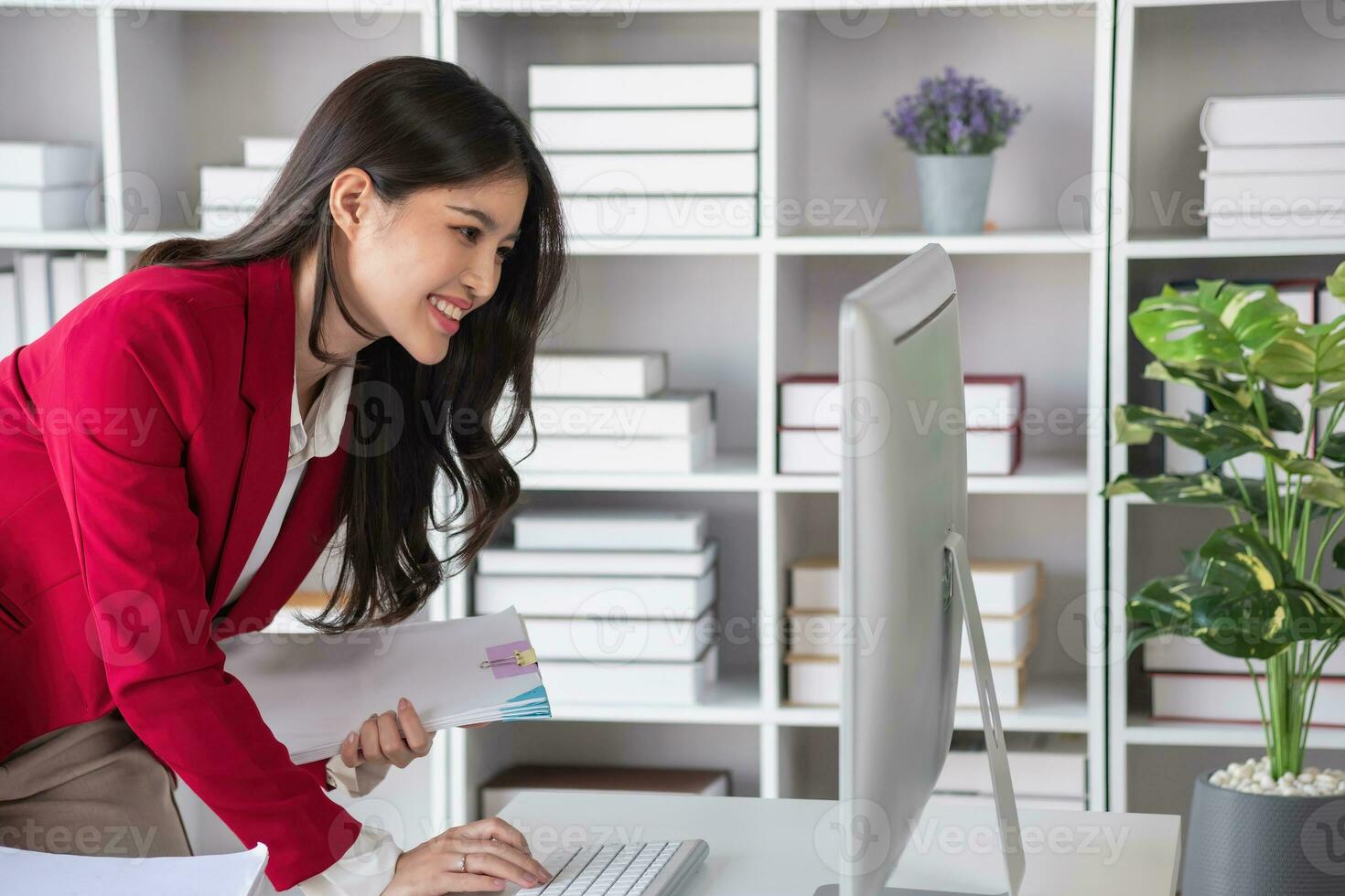 Asian businesswoman in red suit calculating company sales with calculator, laptop, and tablet on table Interior of businesswoman office at the modern workstation. photo