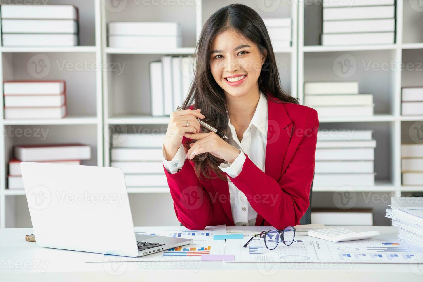Asian businesswoman in red suit calculating company sales with calculator, laptop, and tablet on table Interior of businesswoman office at the modern workstation. photo