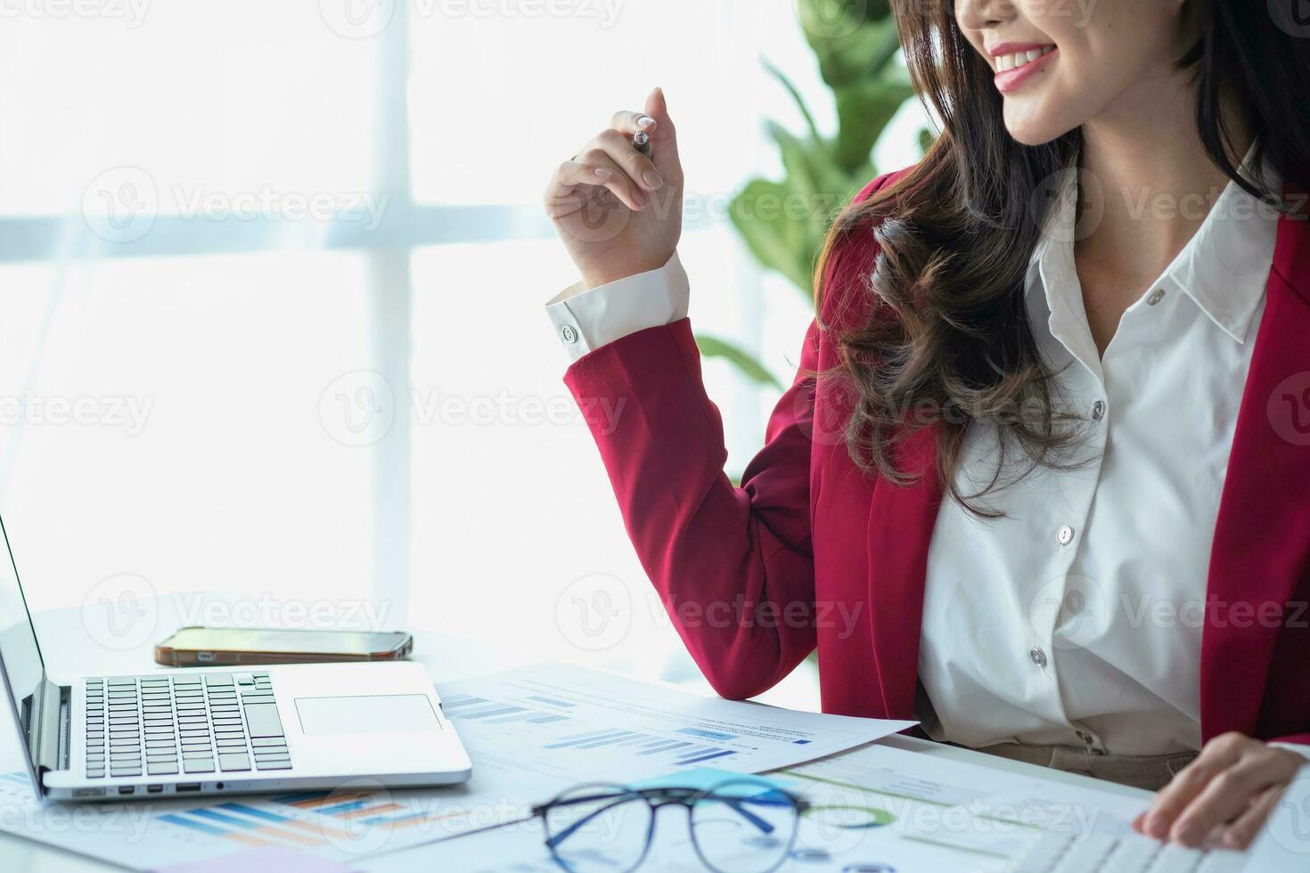 Asian businesswoman in red suit calculating company sales with calculator, laptop, and tablet on table Interior of businesswoman office at the modern workstation. photo