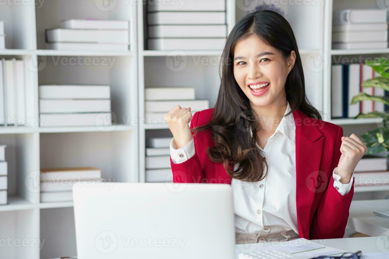 contento asiático mujer de negocios en rojo traje demostración contento gesto participación teléfono inteligente a escritorio retrato de hermosa sonriente mujer de negocios trabajando a un moderno puesto de trabajo. foto