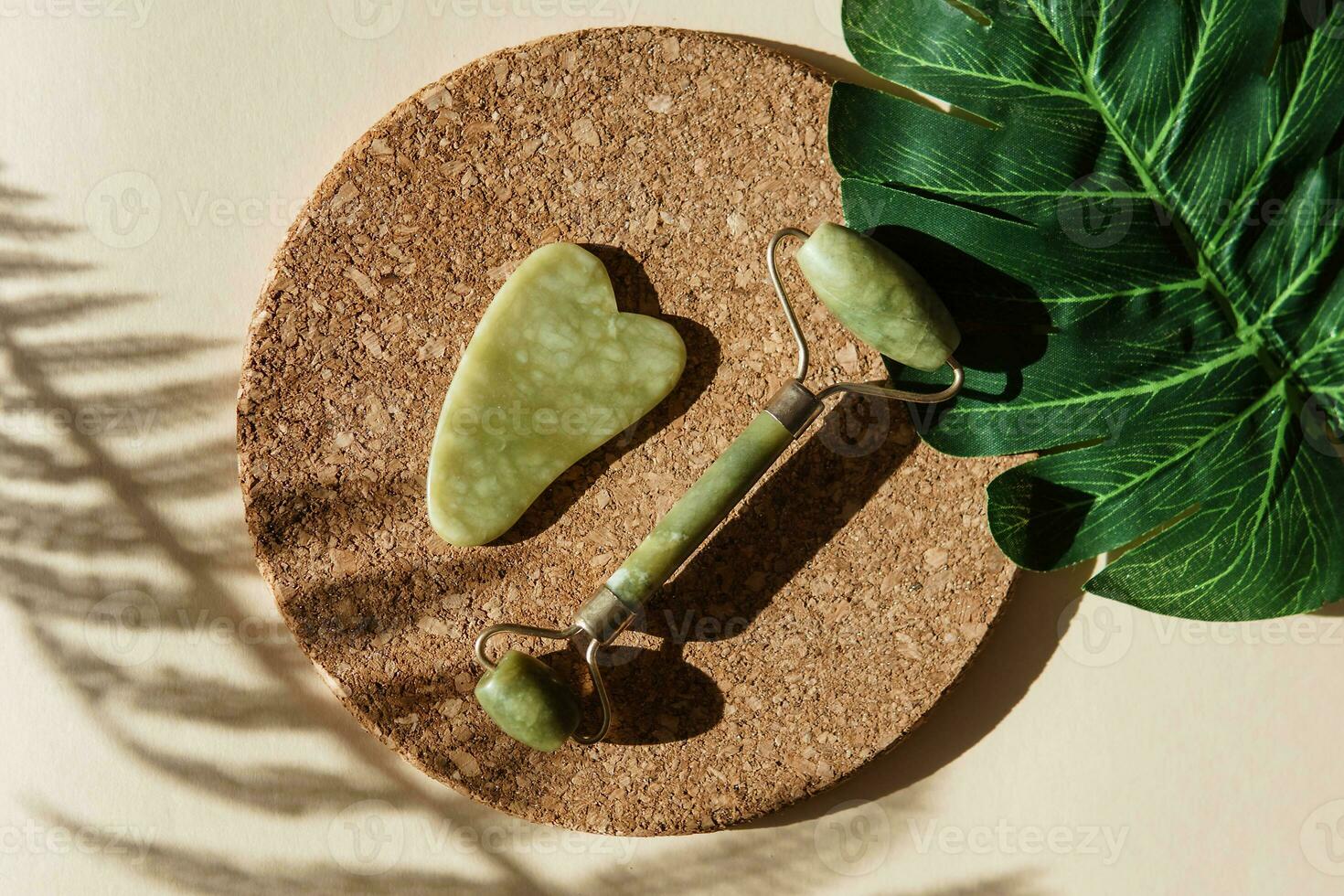 Jade Gua sha scraper and face roller massager on a cork round stand with a monstera leaf. photo