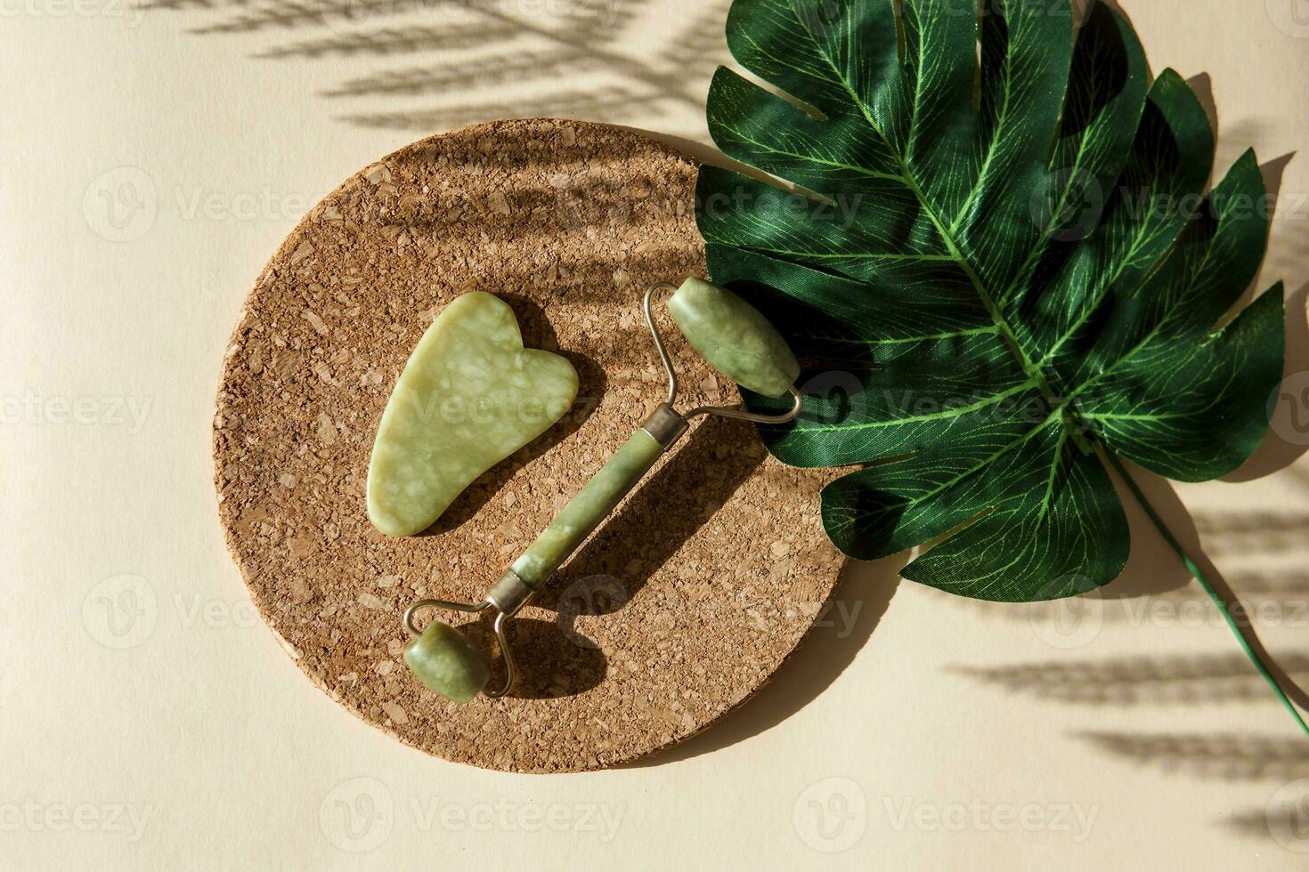 Jade Gua sha scraper and face roller massager on a cork round stand with a monstera leaf. photo