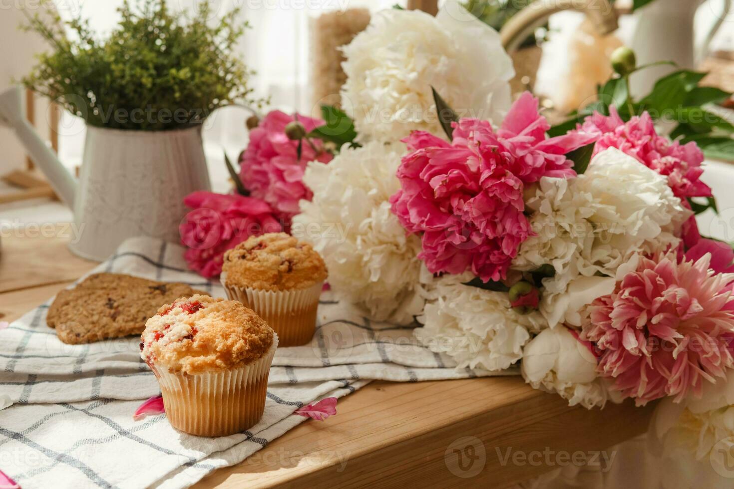 The kitchen countertop is decorated with peonies. The interior is decorated with spring flowers. Pink peonies and sweet cupcakes on a wooden countertop. Interior details. photo