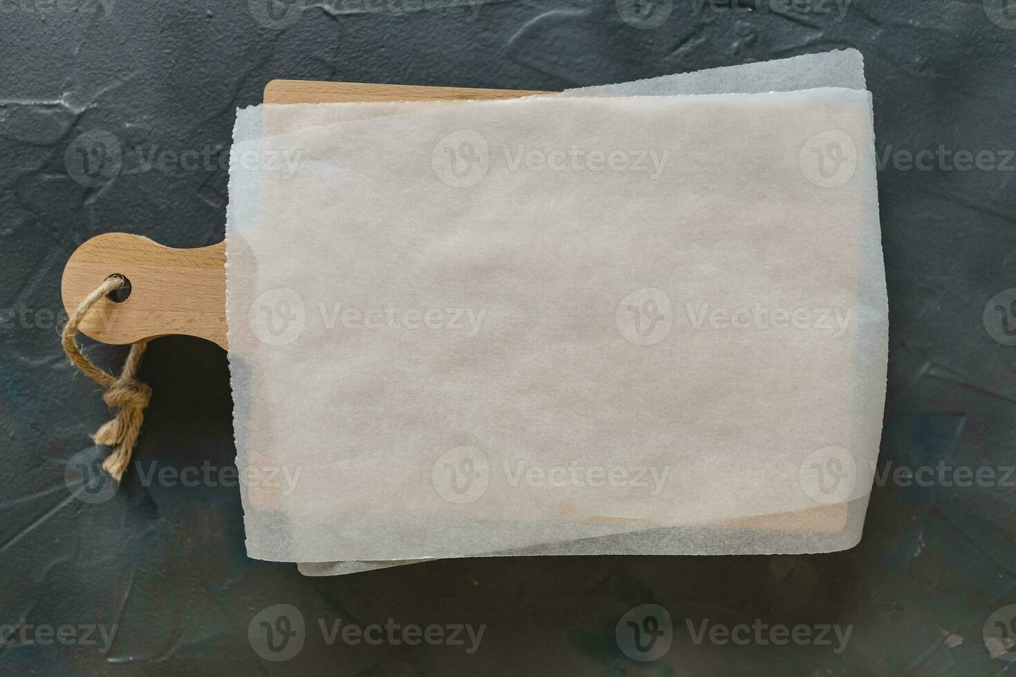 Wooden cutting board and white parchment paper on dark gray background. Mockup for recipes, shopping list or food photo