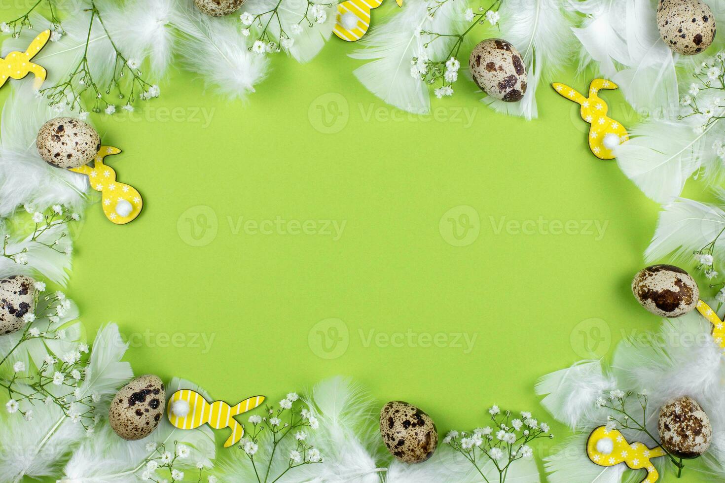 Easter border flat lay with quail eggs, spring flowers, yellow bunnies and white feathers on a green background. Top view. Copy space. photo
