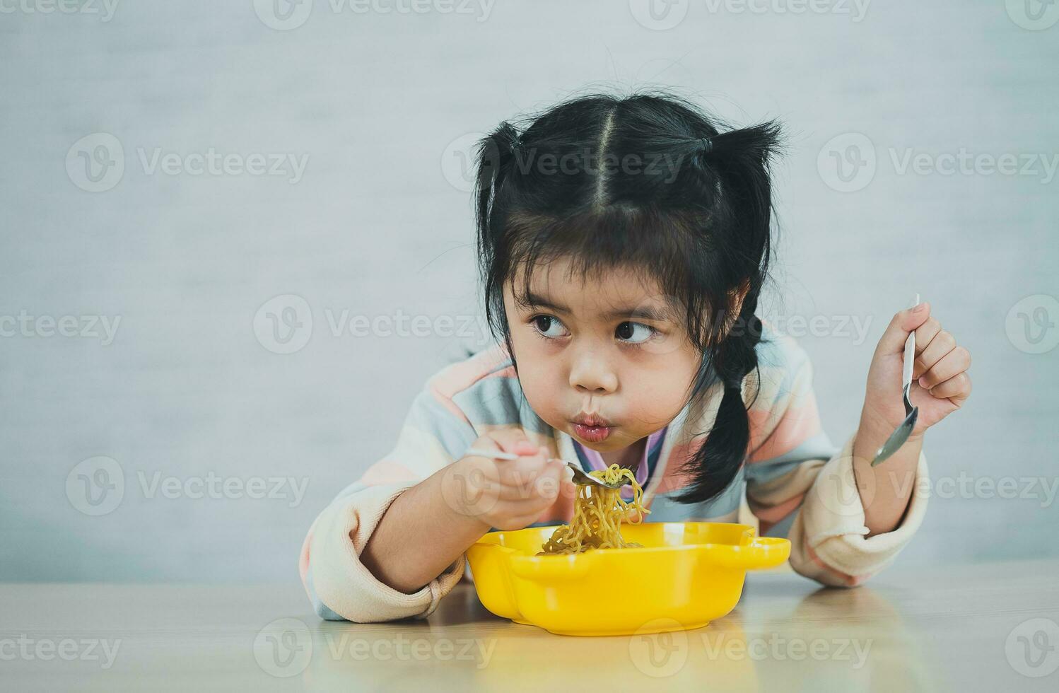 la niña asiática disfruta feliz usando cubiertos, cuchara y tenedor comiendo deliciosos fideos en la cocina en la mesa del comedor. una niña asiática feliz practica comer sola en la mesa del comedor. concepto de comida para bebés foto