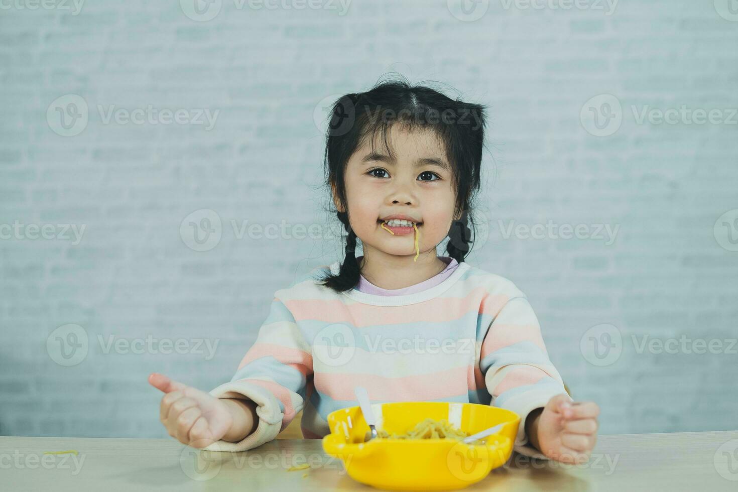la niña asiática disfruta feliz usando cubiertos, cuchara y tenedor comiendo deliciosos fideos en la cocina en la mesa del comedor. una niña asiática feliz practica comer sola en la mesa del comedor. concepto de comida para bebés foto