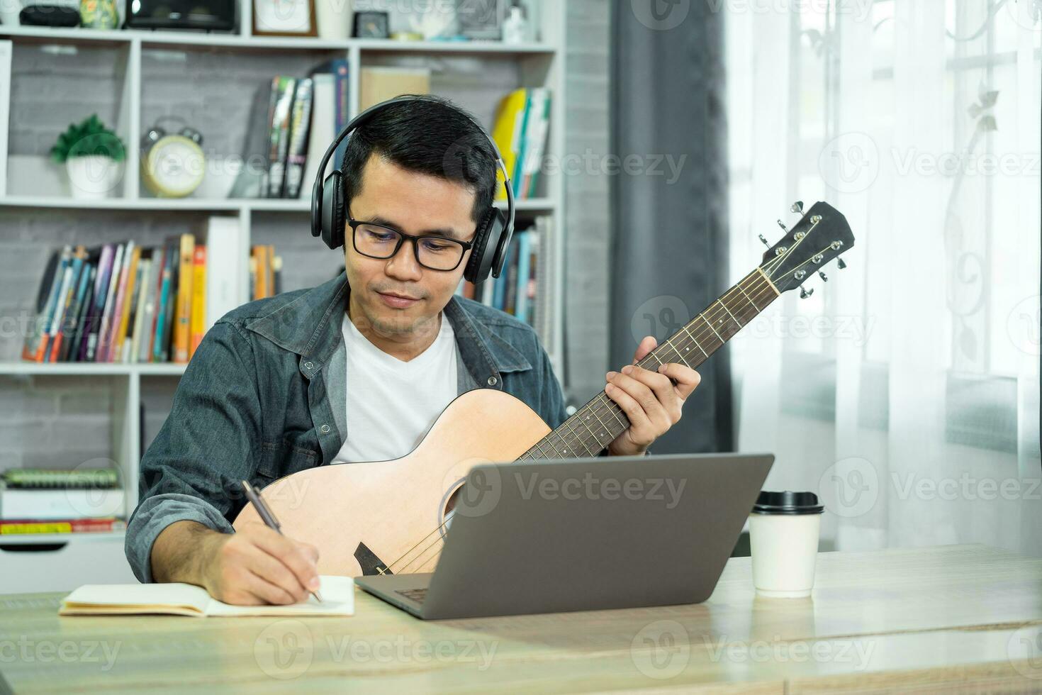 Asian man wearing headphone play guitar and sing a song and learning online with laptop and notesbook and compose writing song, record sound and use computer recording music program. Musician concept. photo