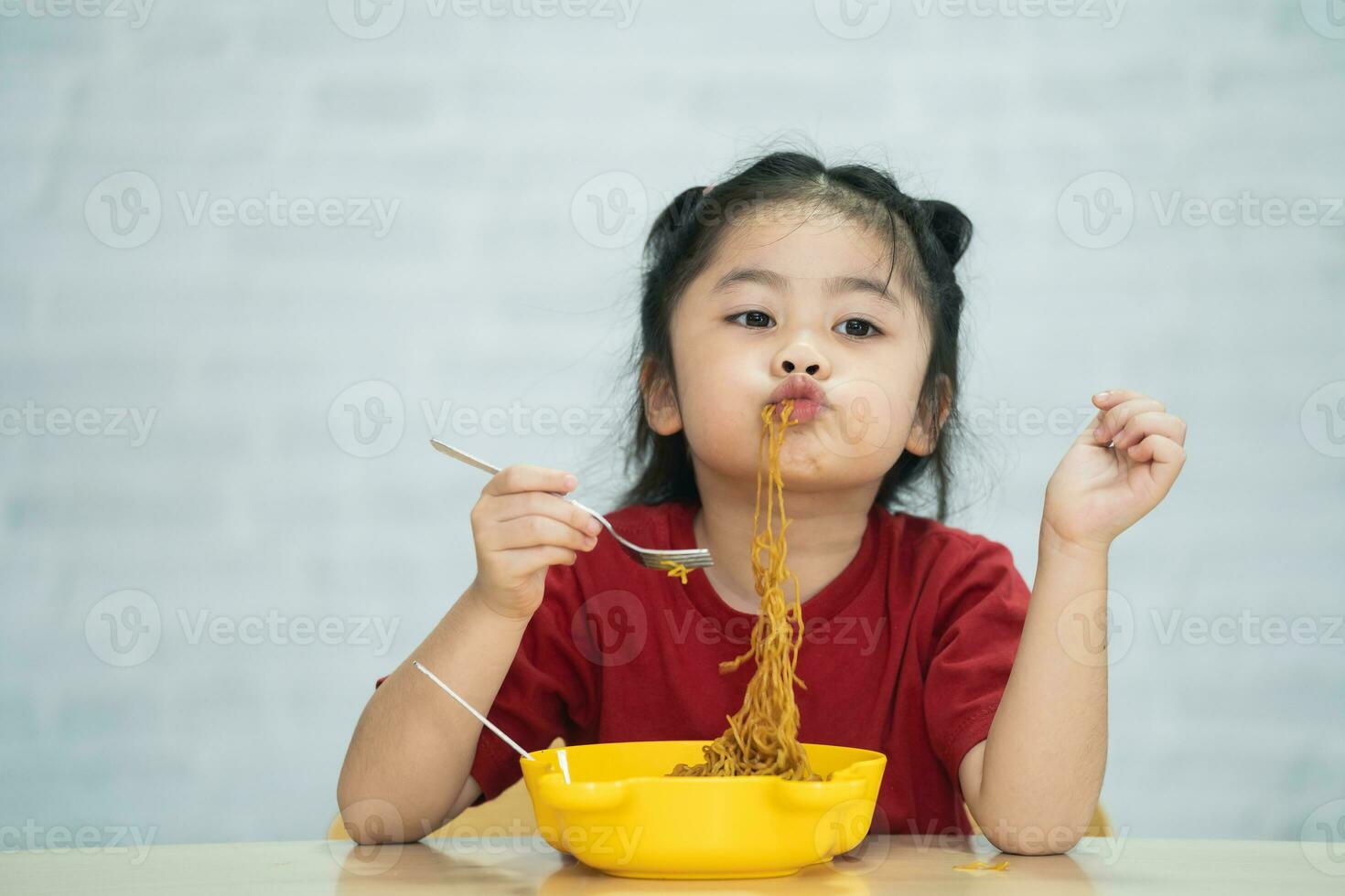 la niña asiática disfruta feliz usando cubiertos, cuchara y tenedor comiendo deliciosos fideos en la cocina en la mesa del comedor. una niña asiática feliz practica comer sola en la mesa del comedor. concepto de comida para bebés foto