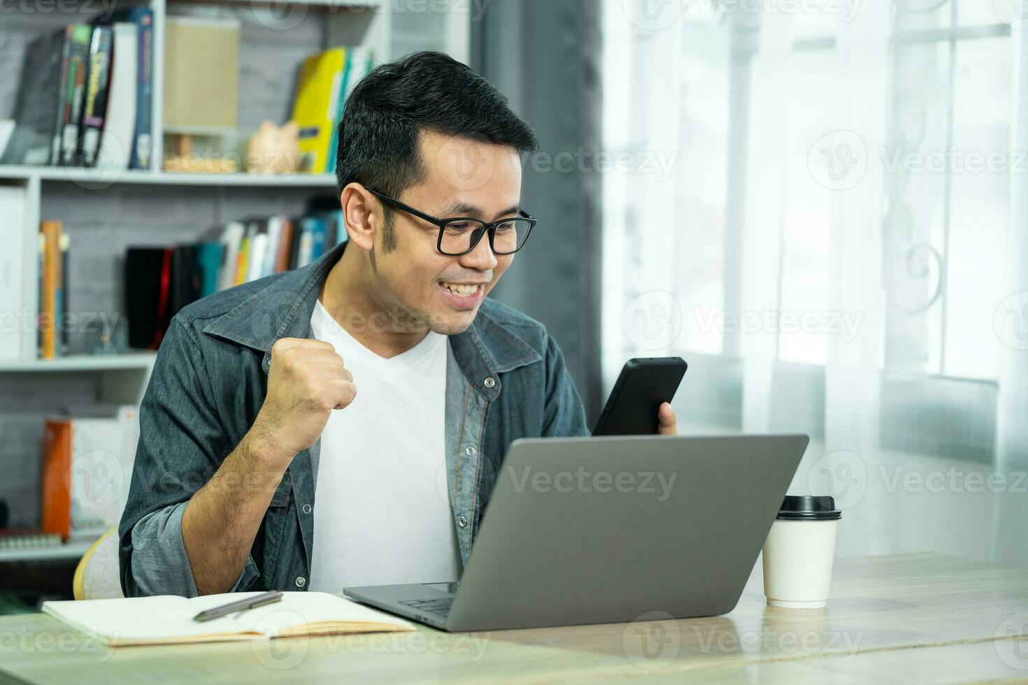 freelance man in eyewear working with laptop and mobile successful cheerful, sitting at wooden table. Pleasant happy man communicating in social network, conferrence vdo call. photo