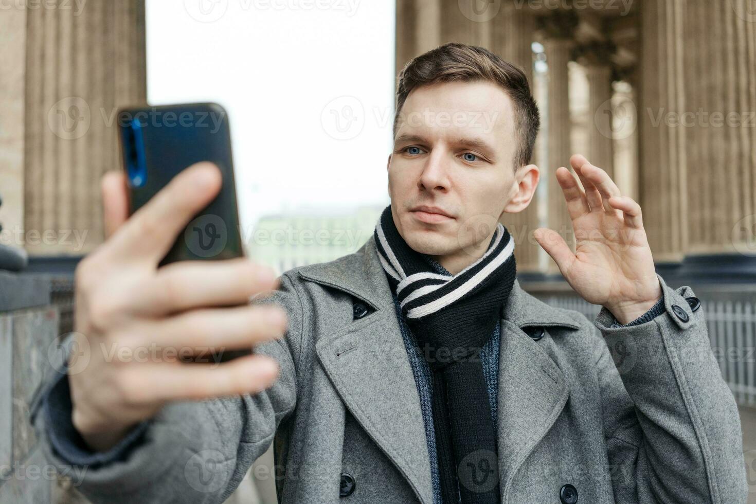 Handsome young man in coat walking on street holds mobile phone and shoots photo