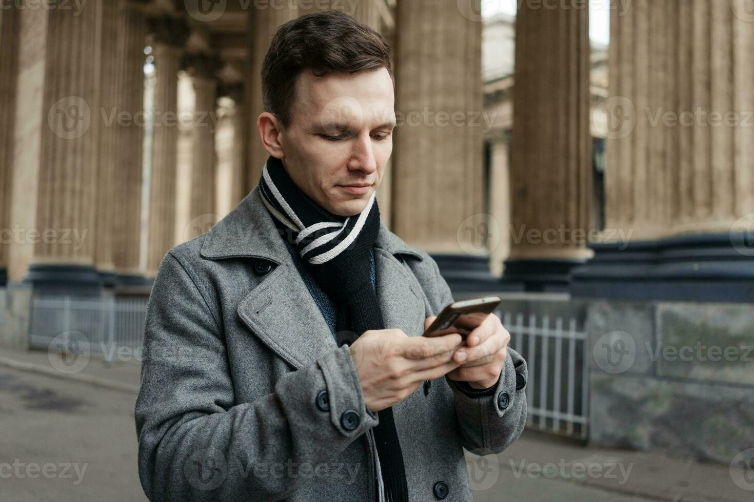 Smiling young man in gray coat is holding scroll text messages in his mobile photo