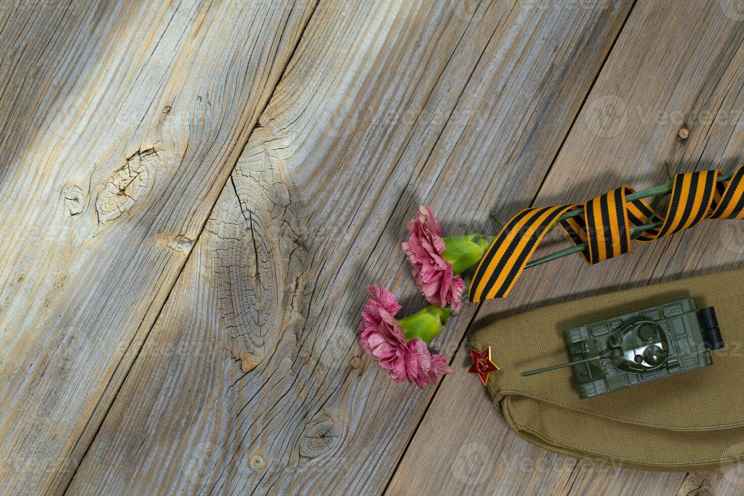 Two pink carnations, Saint George ribbon, tank, and military cap on a wooden surface. photo