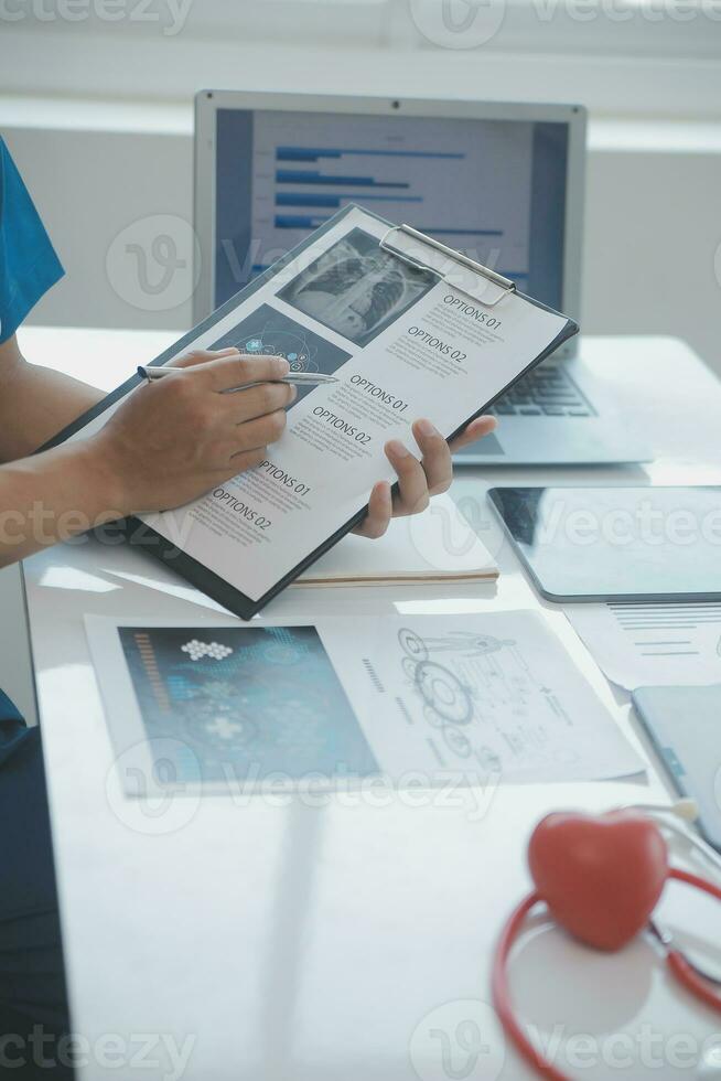 Multiracial medical team having a meeting with doctors in white lab coats and surgical scrubs seated at a table discussing a patients records photo