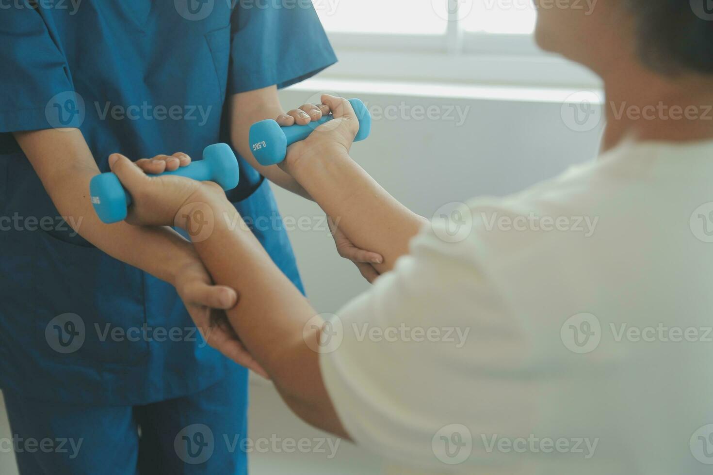 Old woman training with physiotherapist using dumbbells at home. Therapist assisting senior woman with exercises in nursing home. Elderly patient using dumbbells with outstretched arms. photo