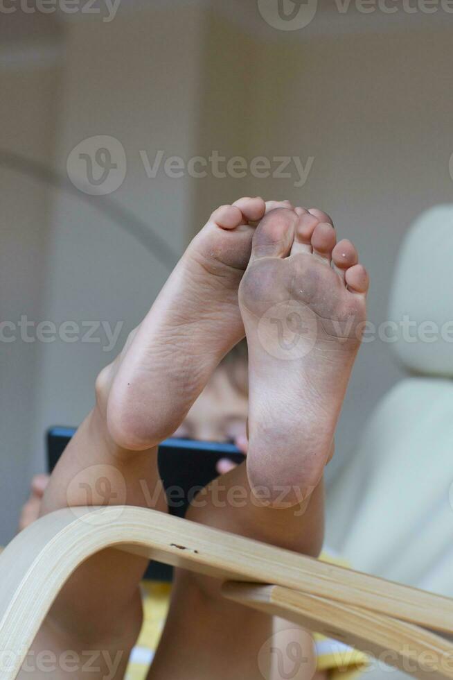 Dirty feet of a  child sitting in the arm chair with his tablet. photo