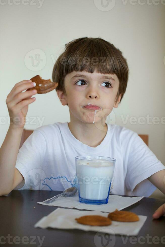 chico de seis años es tomando un vaso de calentar Leche con avena galletas. foto