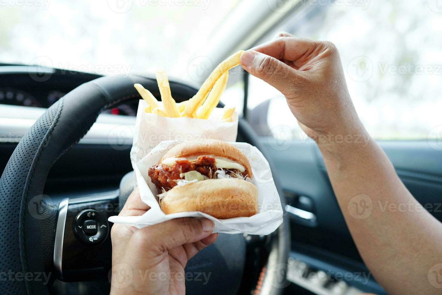 Asian woman driver hold and eat hamburger and french fries in car, dangerous and risk an accident. photo