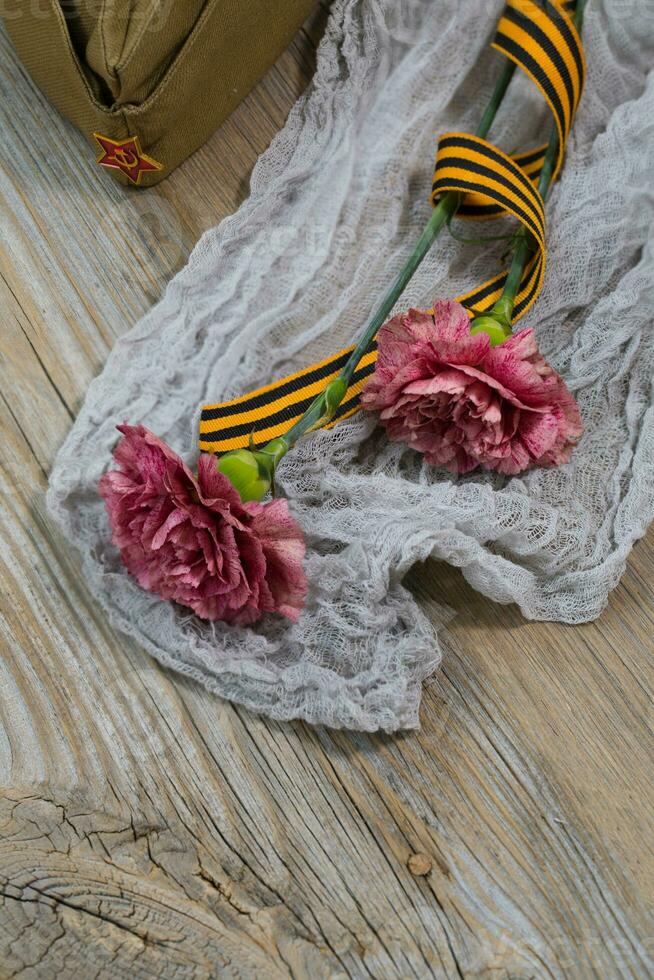 Two pink carnations, Saint George ribbon and military cap on a wooden surface. photo