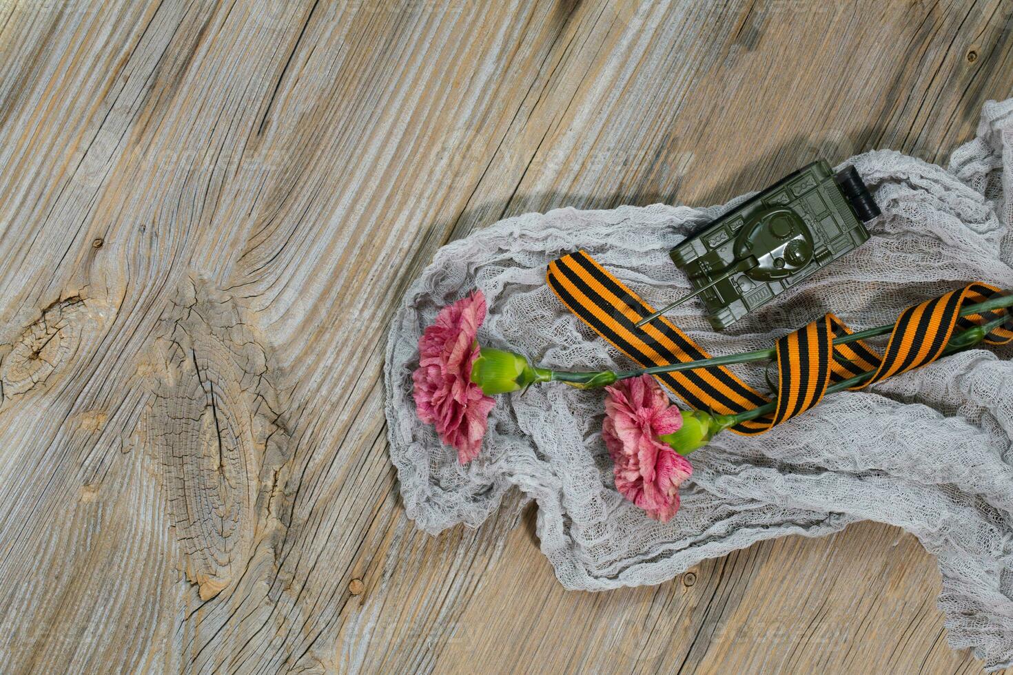 Two pink carnations, Saint George ribbon and military tank on a wooden surface. photo