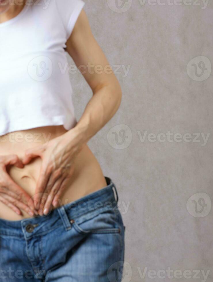 Young lady dressed in jeans shows a heart close to the belly. Closeup photo