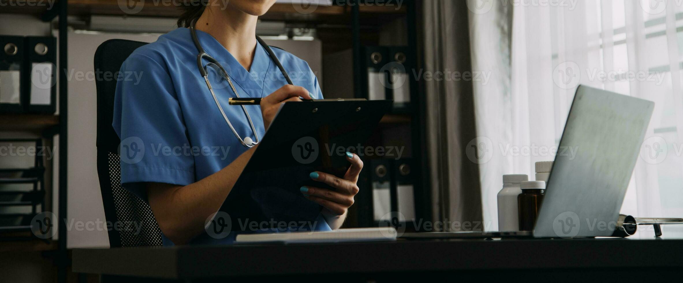 grave hembra médico utilizando ordenador portátil y escritura notas en médico diario sentado a escritorio. joven mujer profesional médico médico vistiendo blanco Saco y estetoscopio trabajando en computadora a lugar de trabajo. foto