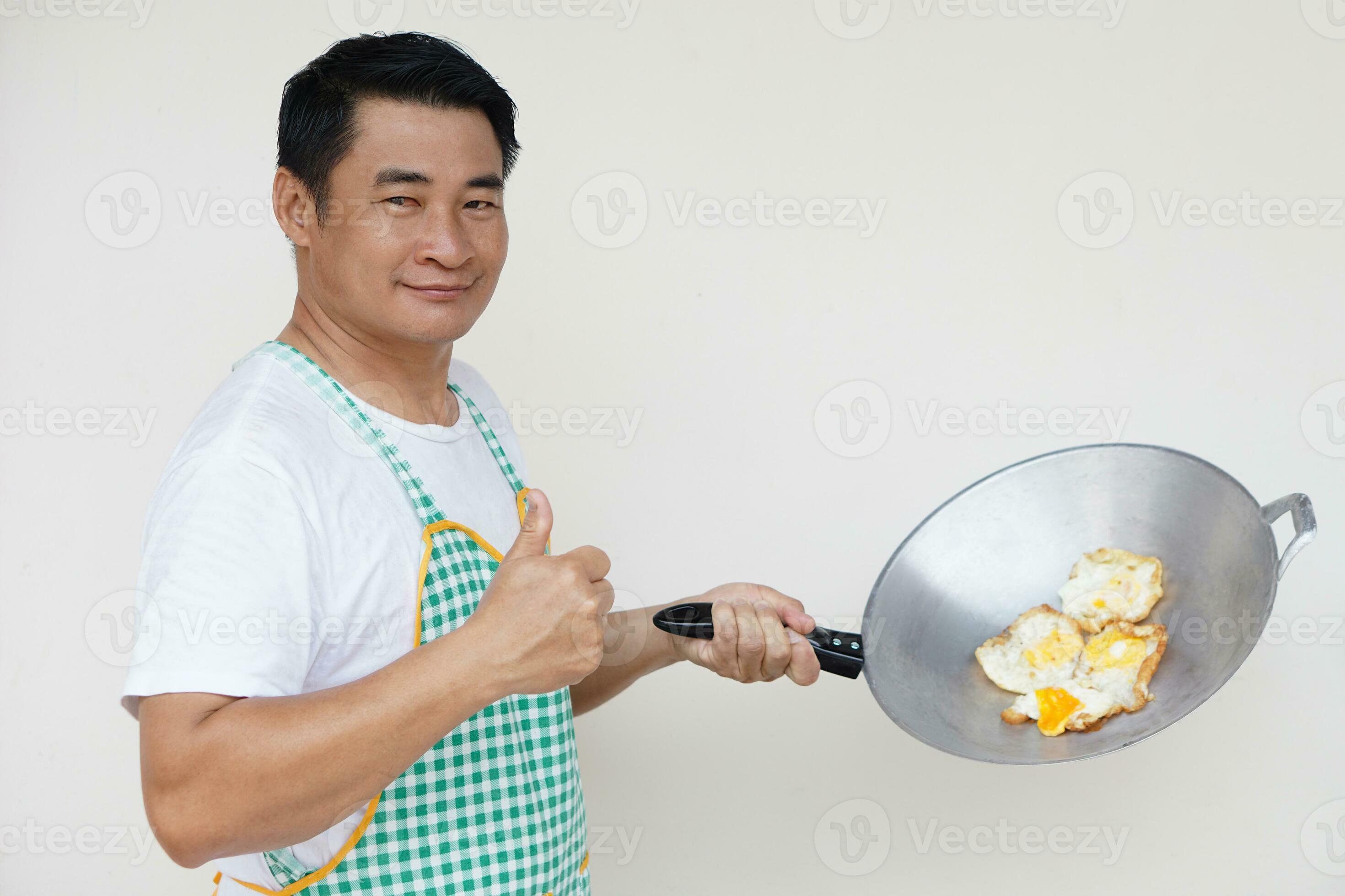 Handsome Asian man is cooking fried eggs, wears white shirt and apron