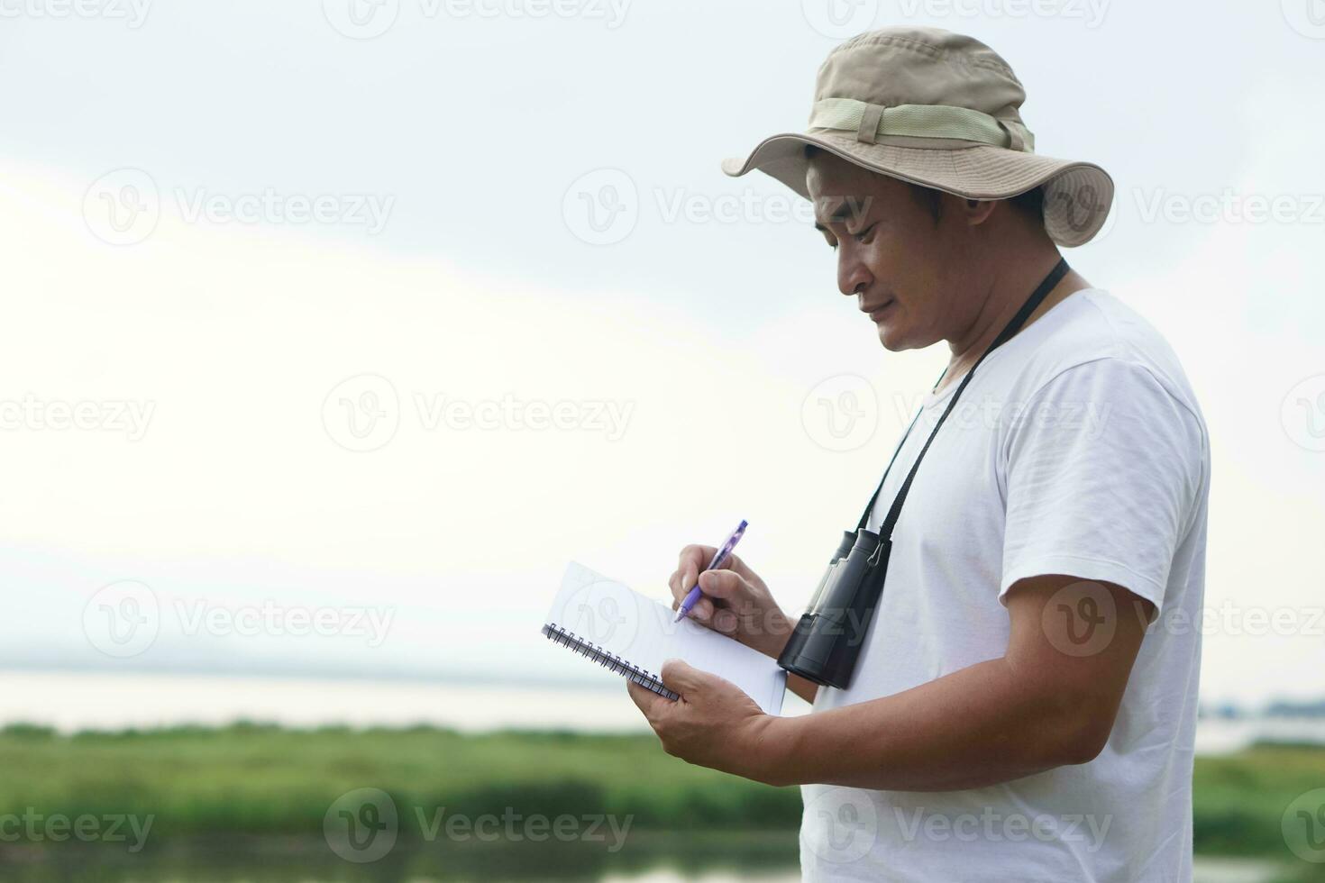 Asian man ecologist is surveying nature at the lake, write on paper notebook. Concept, nature exploration. Ecology study.  Pastime activity, lifestyle. Man explore environment photo