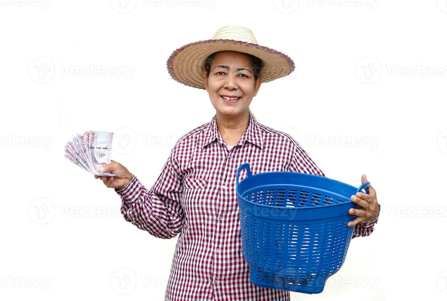 Happy senior woman farmer wears hat, plaid shirt, holds Thai banknotes money and blue basket, isolated on white background. Concept, Farmer happy to get profit, income from doing agriculture. photo