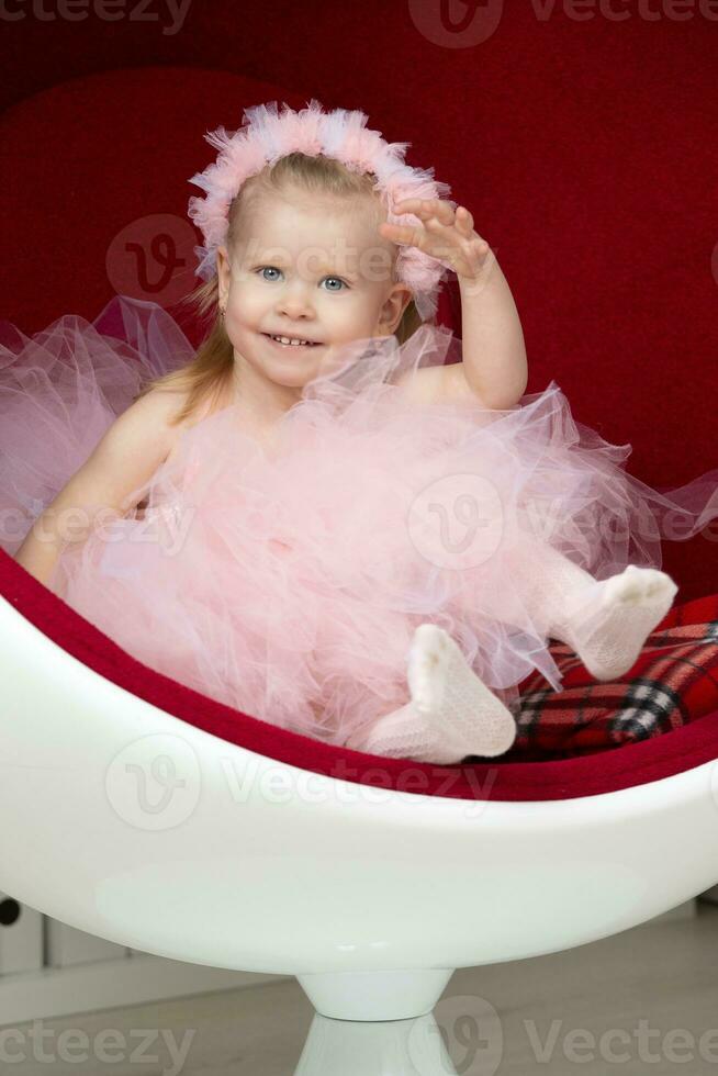 Two-year-old child. A happy little girl in a pink fluffy dress sits on a red chair. photo