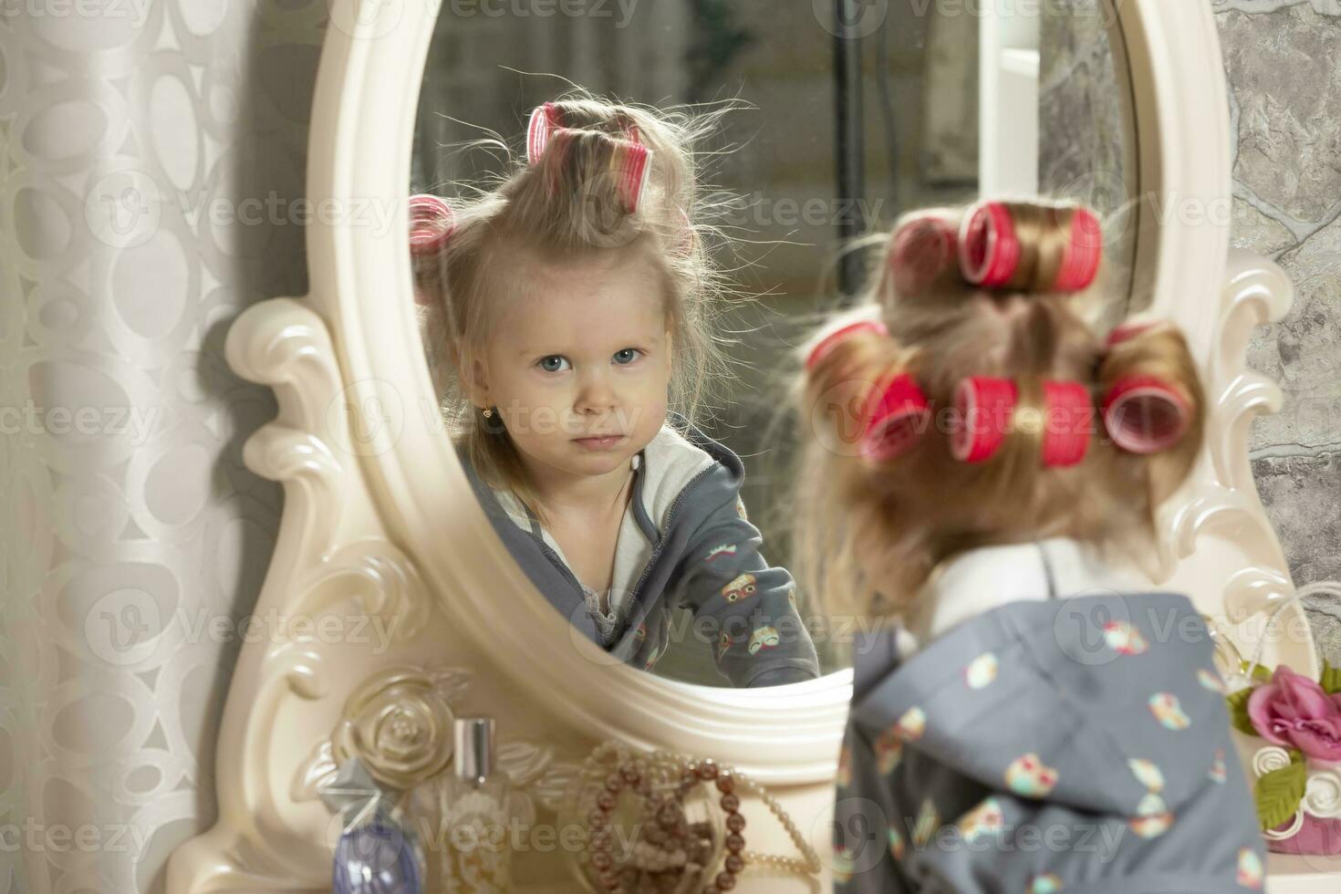 Two-year-old girl. A little girl in curlers looks in the mirror at the dressing table. Party preparation. photo