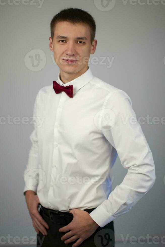 Classic portrait of a young man on a gray background. A guy of about twenty in a white shirt and bow tie. photo