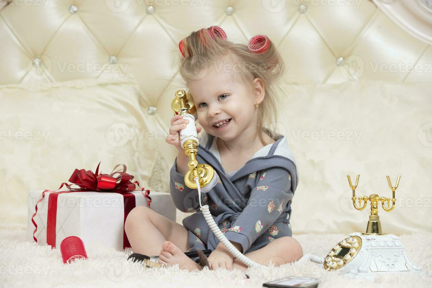 take congratulations on your birthday. A little girl in curlers sits on the bed in the bedroom with a phone. Party preparation. Funny child with a retro telephone. photo