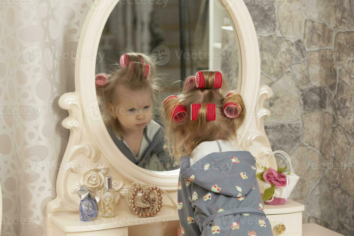 Two-year-old girl. A little girl in curlers looks in the mirror at the dressing table. Party preparation. photo