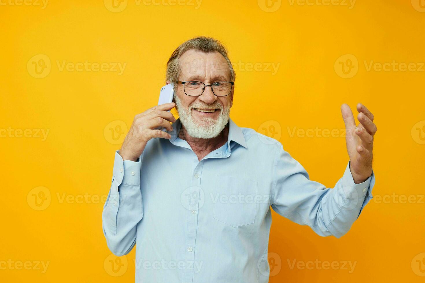 Photo of retired old man in a blue shirt and glasses talking on the phone yellow background