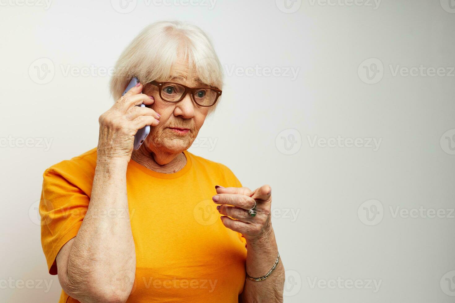 Portrait of an old friendly woman in casual t-shirt glasses talking on the phone light background photo