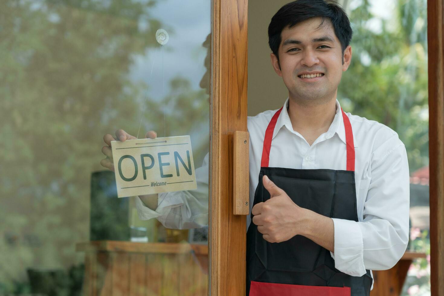 Business owner thumbs up to show his readiness to welcome customers. waitress man turning on open sign board on glass door in modern cafe coffee shop. New normal, small business, cafe, life style. photo