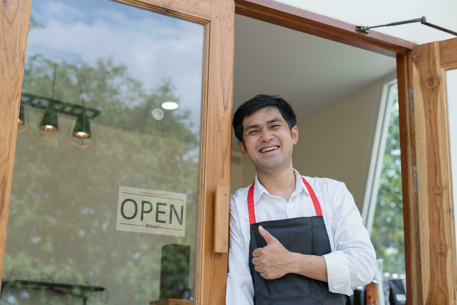 Business owner thumbs up to show his readiness to welcome customers. waitress man turning on open sign board on glass door in modern cafe coffee shop. New normal, small business, cafe, life style. photo