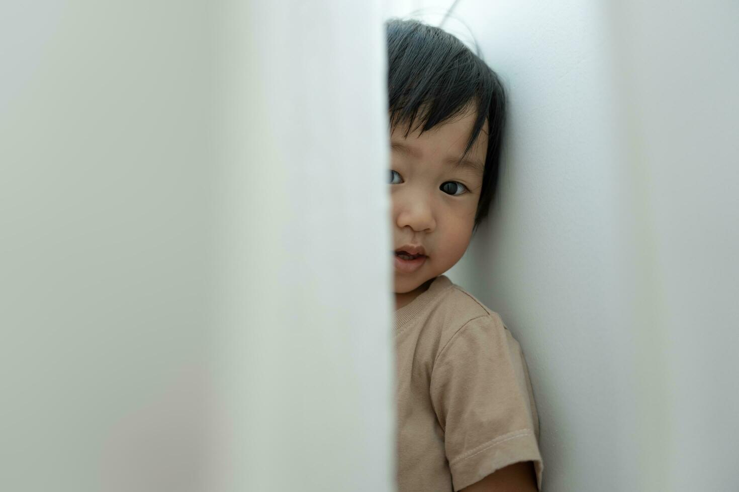 pequeño asiático niños niño ocultación detrás cortinas , jugando esconder y buscar en pie siguiente un pared casa, adorable niño teniendo divertido en el hogar. contento familia tener divertido con niños, actividad, aprendizaje foto