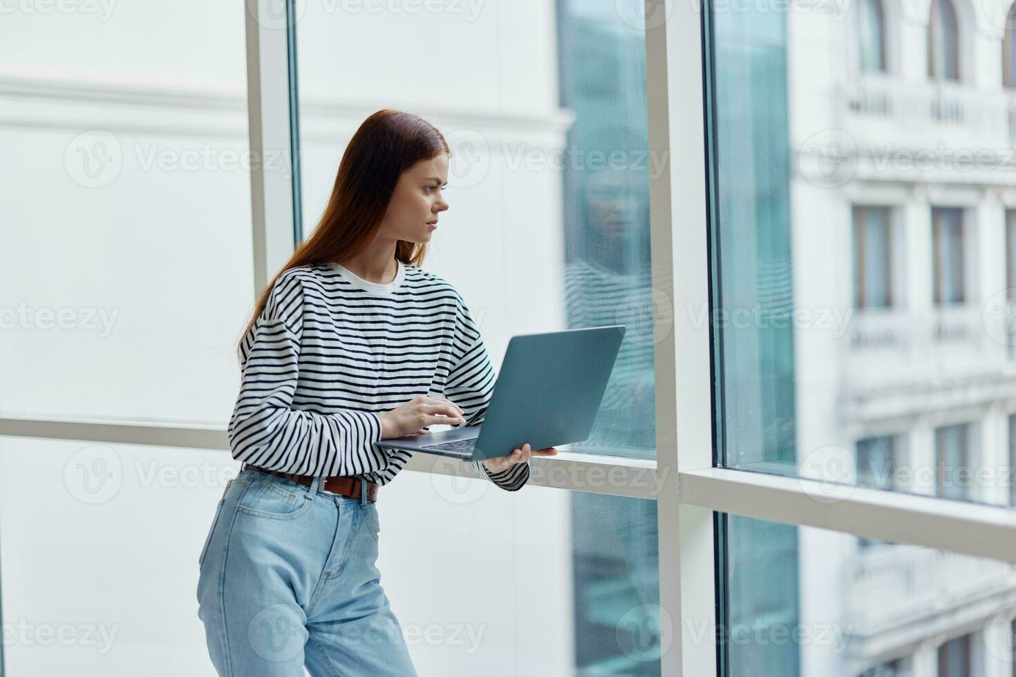 A woman standing near a large window in the city and holding a laptop in her hand looking out the window at the cityscape, a copy space photo