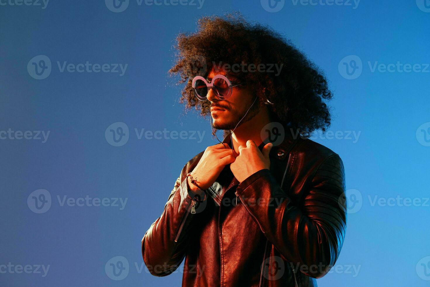 Portrait of a stylish man with curly hair with glasses and headphones on a blue background multinational, colored light, black leather jacket trend, modern concept. photo