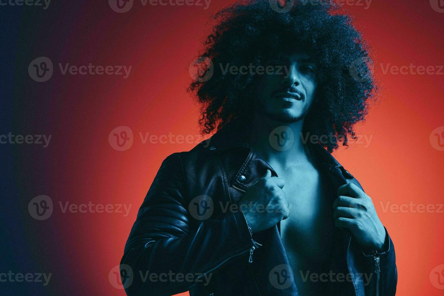 Portrait of fashion man with curly hair on red background multinational, colored light, black leather jacket trend, modern concept. photo