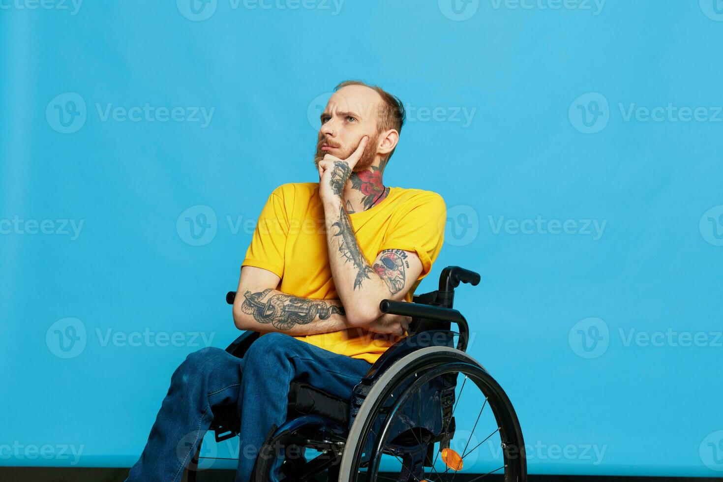 a man sits in a wheelchair thoughtfulness in a t-shirt on a blue background in the studio, the concept of a free barrier-free environment for people with disabilities photo